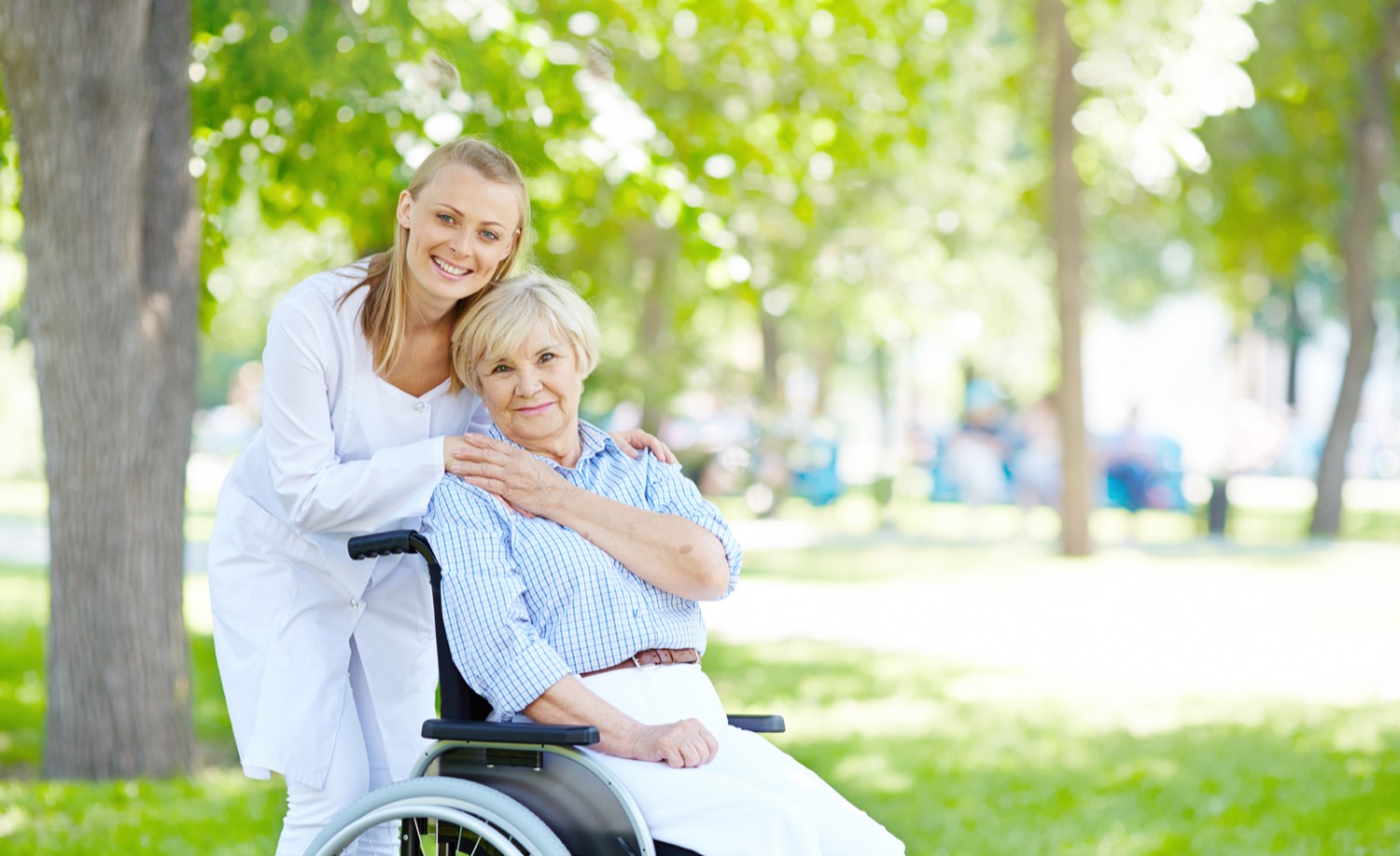 A caregiver standing beside an older woman outdoors, both smiling